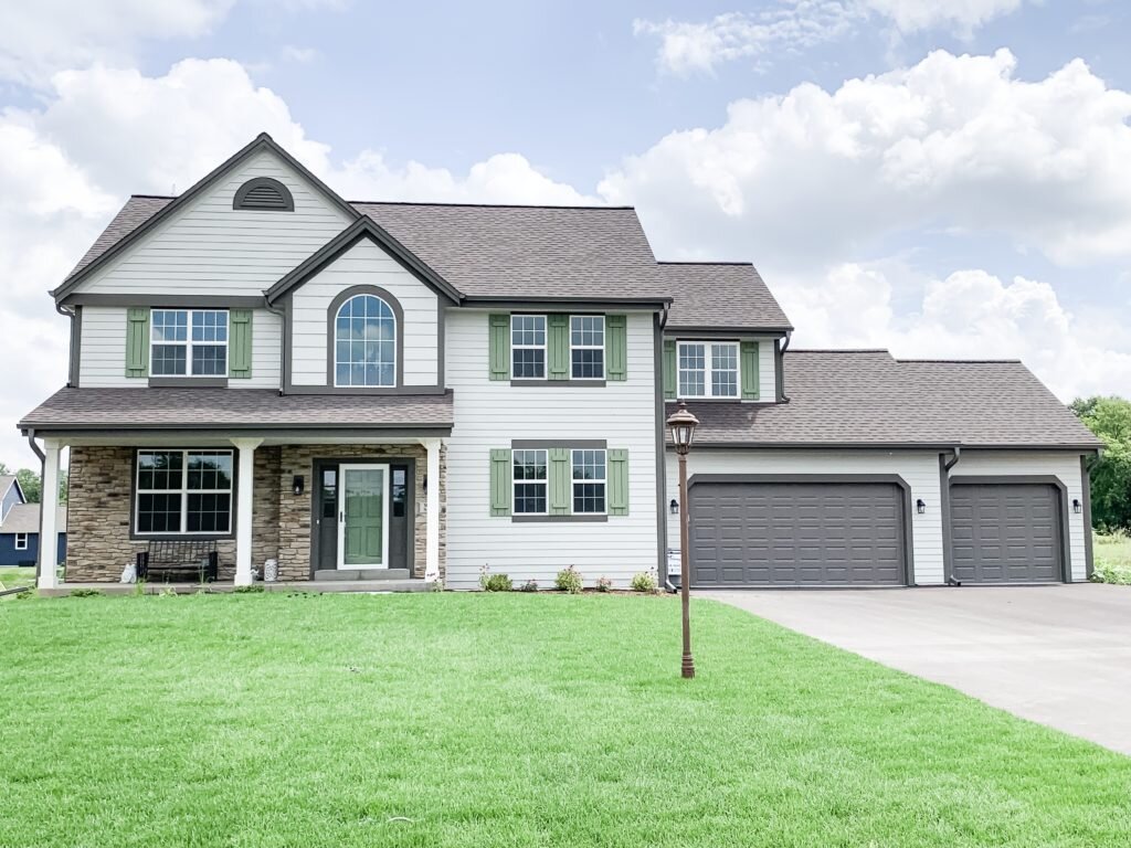 A large, two-story family home with a symmetrical facade, featuring stone and siding accents. The home has green shutters and a three-car garage, with a well-manicured lawn and a charming lamp post in the front yard.