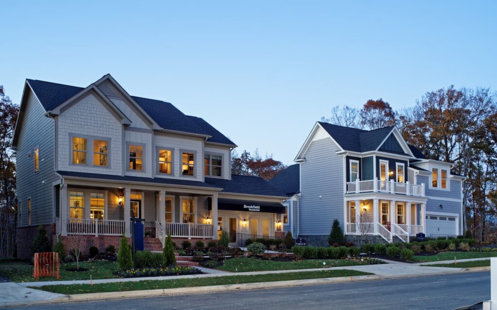 Two side-by-side modern houses in a suburban neighborhood, illuminated warmly in the evening. Both homes feature large windows, porches, and beautifully landscaped front yards, providing a cozy and inviting atmosphere. The sky is a clear evening blue.