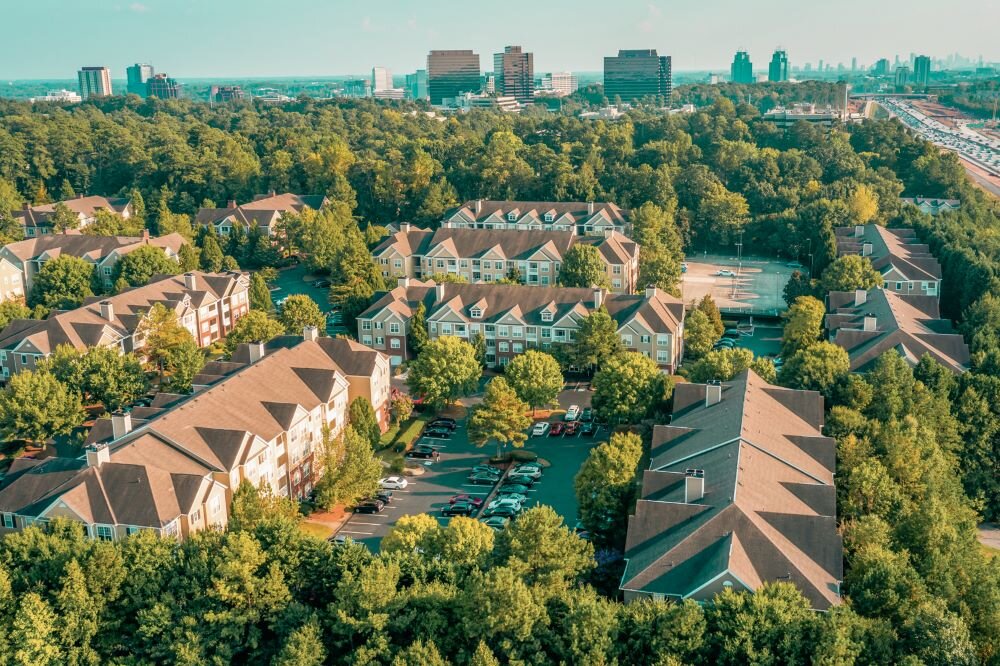 Aerial view of a large residential apartment complex surrounded by lush green trees and a city skyline in the background, highlighting an expansive neighborhood with multiple buildings and well-planned infrastructure.