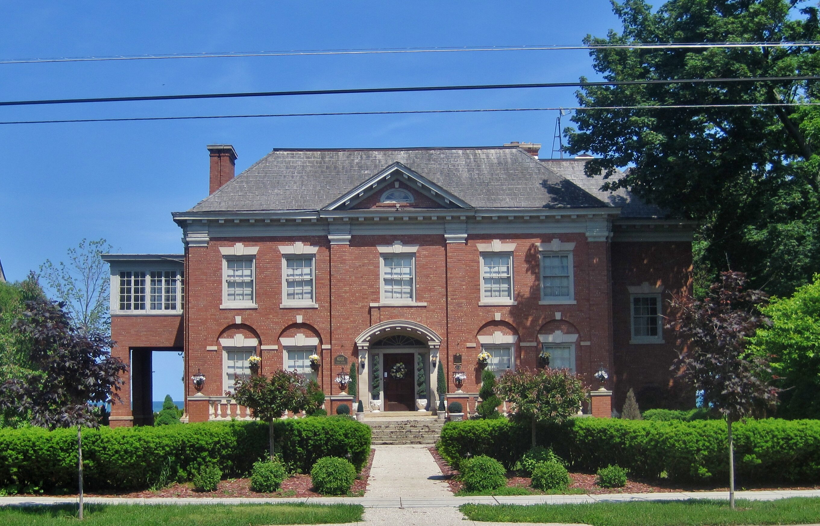 A long row of modern, two-story townhouses painted in soft gray and white tones, with small front yards and young trees lining the street under a bright blue sky.