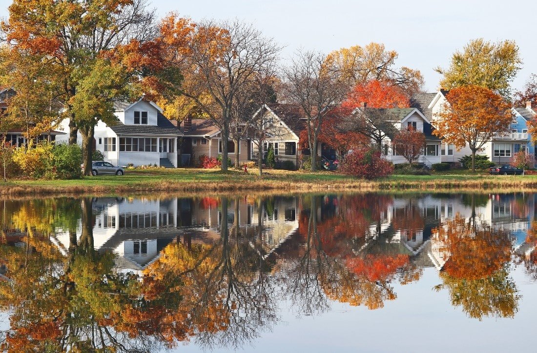 A row of lakeside houses with beautiful autumn foliage reflected in the calm water. The scene captures vibrant colors of red, orange, and yellow leaves, showcasing the picturesque beauty of a tranquil residential neighborhood in fall.