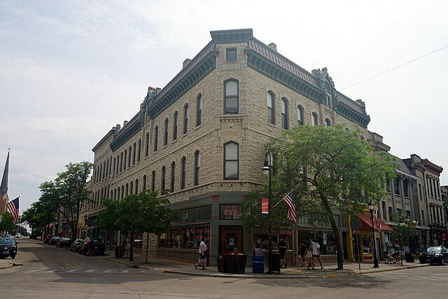 A large, historic stone building located at a street corner, surrounded by small shops and pedestrians on a sunny day.