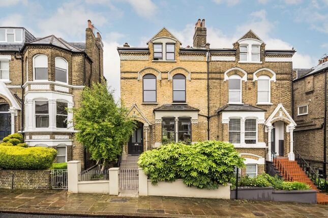 Victorian-style townhouses in a residential area. Each building has a distinct brick facade with ornate detailing, large windows, and decorative greenery along the entrance. The street is lined with lush foliage and has a wet pavement, adding to the historic charm of the architecture.