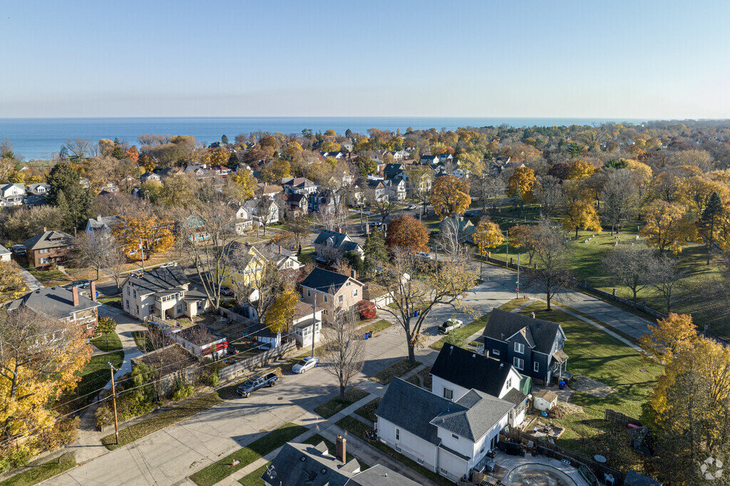  An aerial view of a residential neighborhood with houses surrounded by tall trees showcasing autumn colors. The neighborhood is situated near a large body of water, creating a picturesque scene of suburban living.