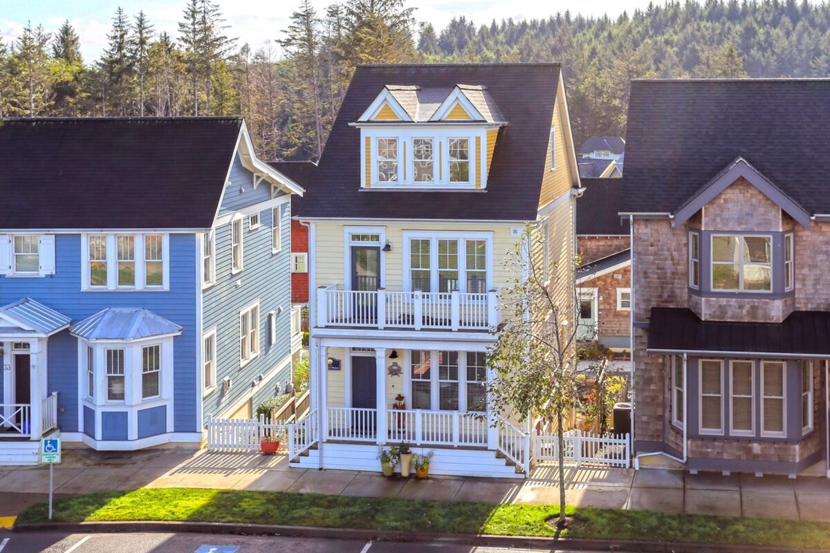 A vibrant, narrow three-story home with a white and yellow exterior, accented by white balconies on each floor. The house is nestled between two similarly styled homes with coastal design features, set against a backdrop of dense forest.