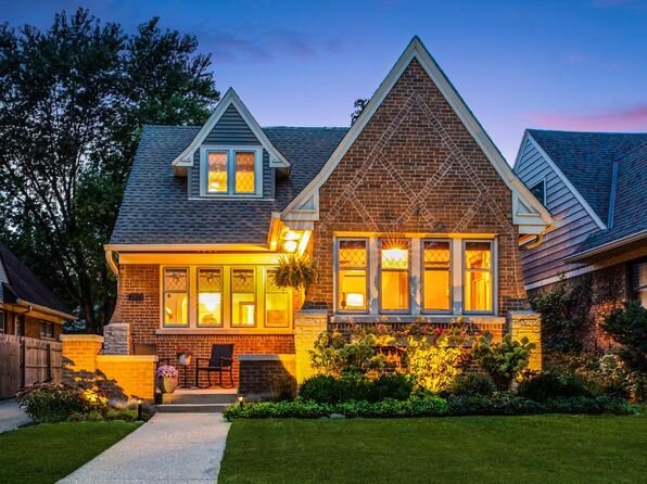 A brick Tudor-style home illuminated warmly at dusk, with glowing interior lights visible through vintage-style windows, a front porch with seating, and neatly maintained landscaping around the front.