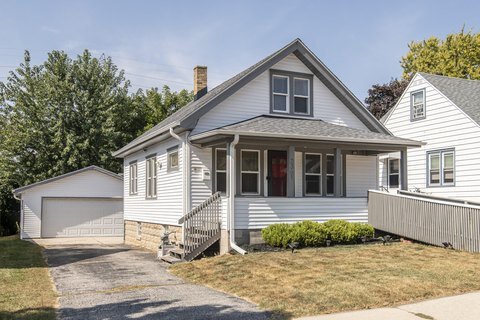 A quaint white house with a covered front porch, sloping rooflines, and a detached garage in the background. The driveway runs along the side, creating a peaceful suburban look.
