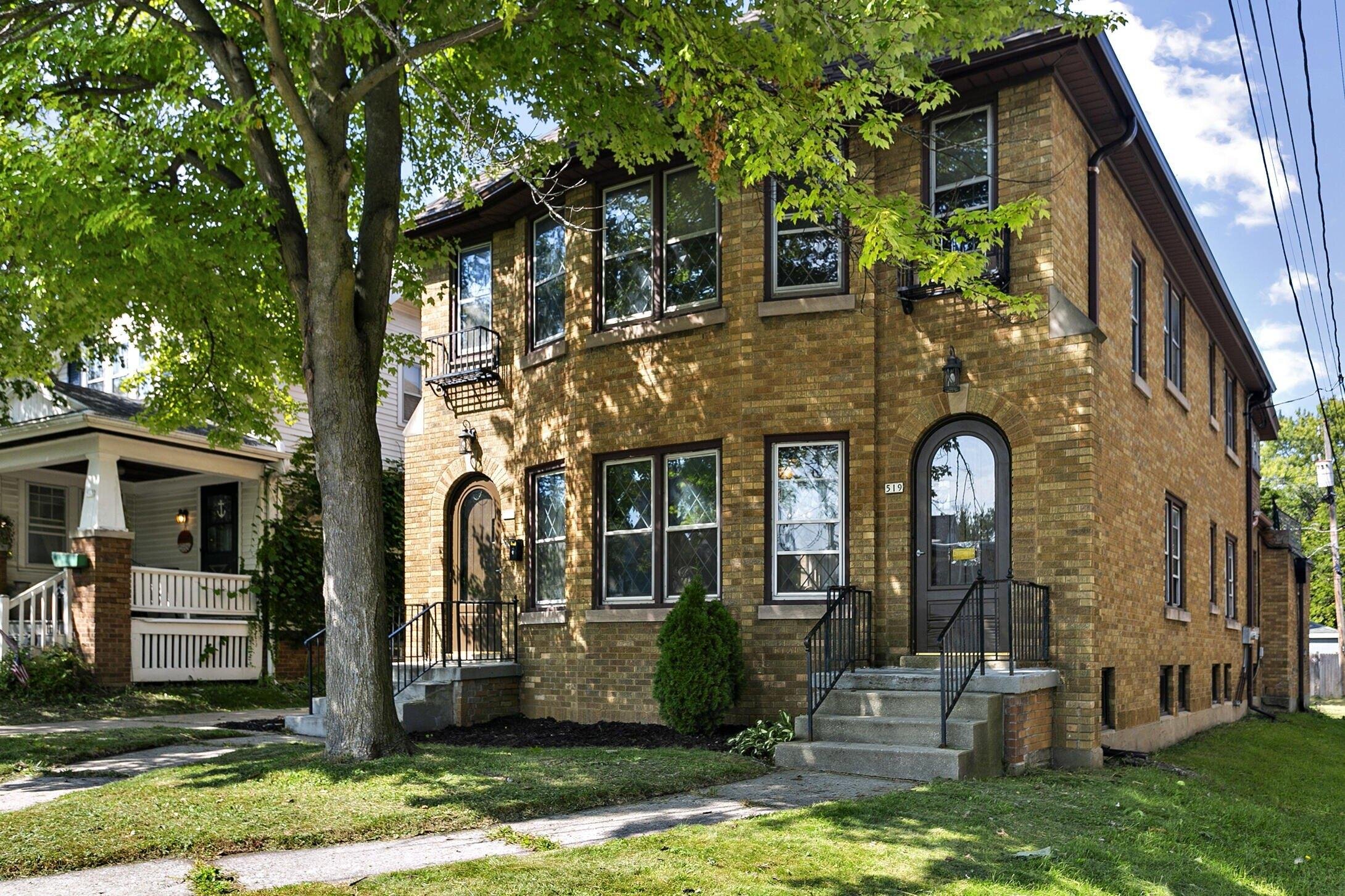 A beautiful two-story brick apartment building with two distinct entryways, both featuring arched doors, surrounded by trees and well-maintained greenery in a cozy neighborhood.