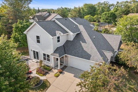 Aerial view of a modern two-story white house with a well-maintained front yard and landscaped flower beds. The home features a gabled roof, spacious driveway, and a cozy porch surrounded by greenery, giving it a welcoming suburban appeal.