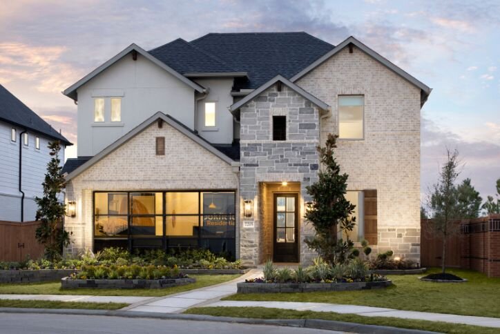 A modern two-story home with a light brick exterior, featuring large windows and a well-maintained front yard. The home includes a mix of stone and siding elements, a prominent front door, and is surrounded by landscaped plants and greenery, set against a dusk sky.