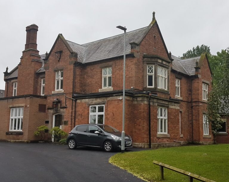 A grand, historic two-story red brick building with Gothic architectural elements. The property includes intricate stone details around the windows, a chimney, and a tall, arched roof, with a car parked in the front on a paved driveway.