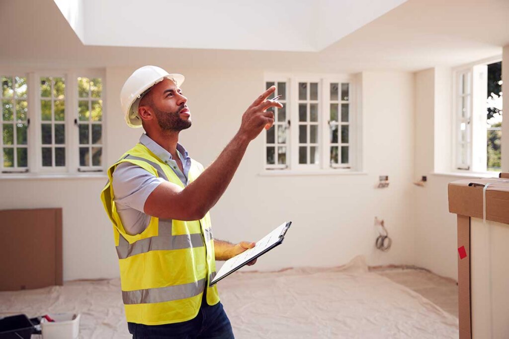 A construction inspector in a bright yellow vest and hard hat, holding a clipboard and pointing at the interior ceiling of a house under renovation. The setting features freshly painted walls and large windows, indicating a newly remodeled property.