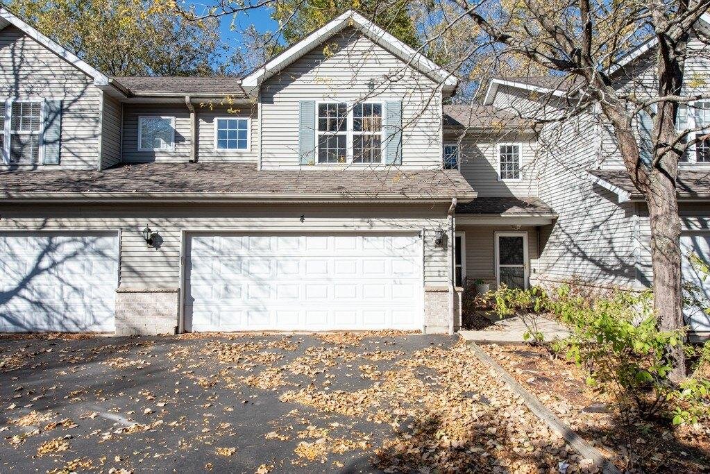 A modern, gray townhouse with two large garage doors in the front, surrounded by trees with fallen autumn leaves scattered on the driveway.