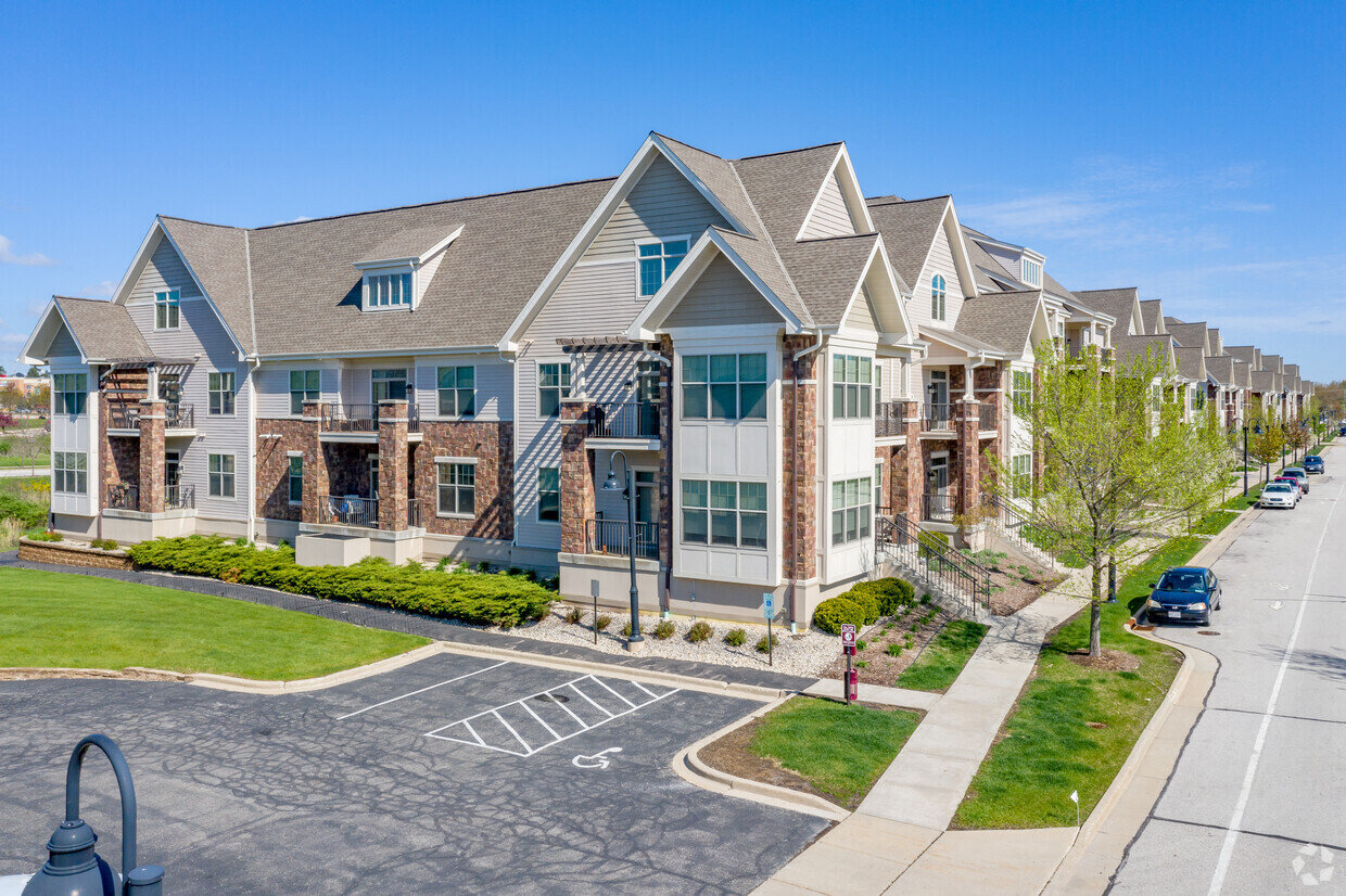 A contemporary apartment complex featuring stone accents and large windows on a clear, sunny day. The building is surrounded by manicured lawns, pathways, and parking spaces, highlighting a well-maintained residential area.