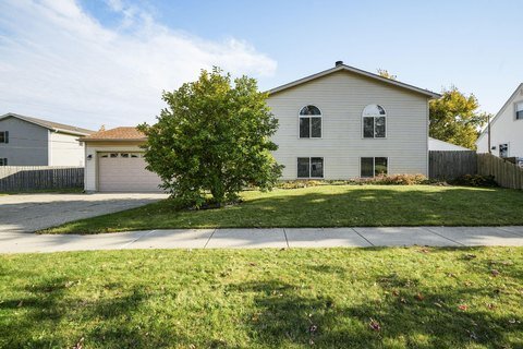 A light beige split-level home with large arched windows on the second floor. The front lawn is wide with a small tree in the center, and a two-car garage is visible to the left side of the house.