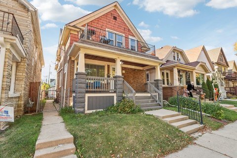 A cozy, red-brick bungalow with a covered porch, dark wooden accents, and decorative stonework. The house has a front staircase leading to the porch, flanked by small gardens and mature trees that frame the property beautifully.