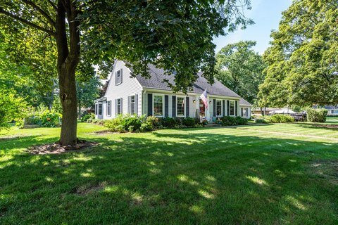 A charming single-story country-style house with white siding and a pitched roof. The house is surrounded by lush, well-maintained green lawn, with large trees providing shade in the background.