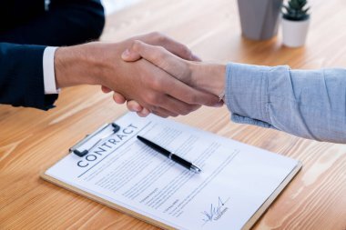 A close-up image showing a handshake between two individuals across a wooden table. The handshake signifies the successful agreement of a contract, visible beneath their hands, which is accompanied by a black pen. The background is slightly blurred, emphasizing the handshake and the contract as the primary focus.