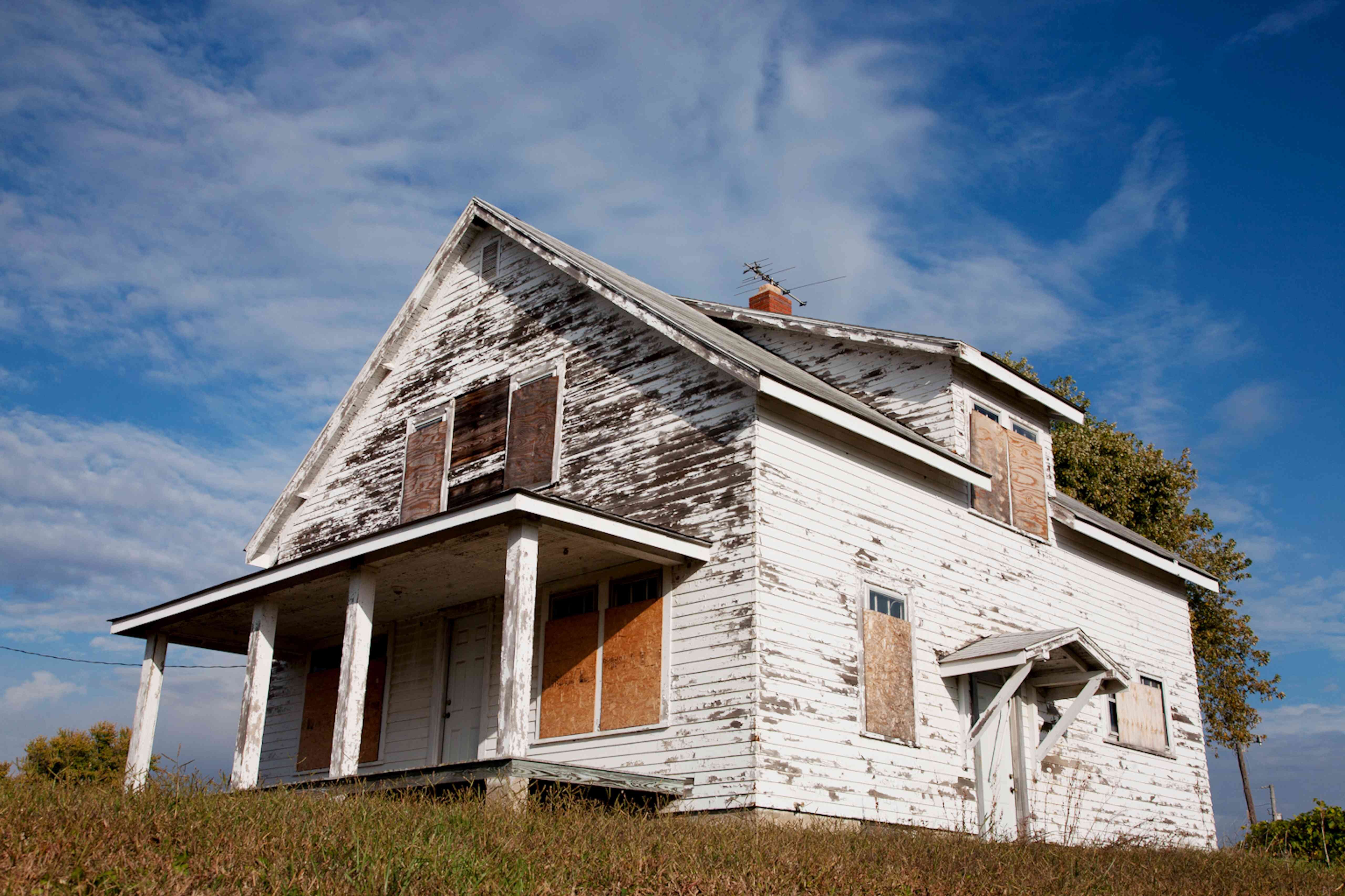 An old, abandoned white house with peeling paint and boarded-up windows sits isolated in a grassy field under a partly cloudy sky. The house appears to be in poor condition, indicating a property that might be vacant or distressed.