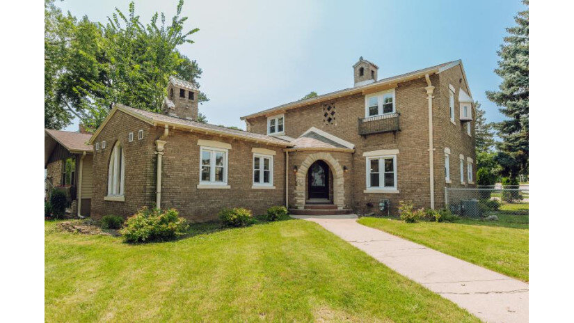 A charming, two-story brick house featuring an arched doorway, vintage architectural details, and a low-maintenance lawn. The extended wing to the side adds character, while the front path and modest landscaping enhance the inviting feel.