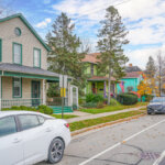 A light gray house with green trim and accents, featuring a covered front porch and a white car parked in front. The neighborhood appears calm and lined with trees.