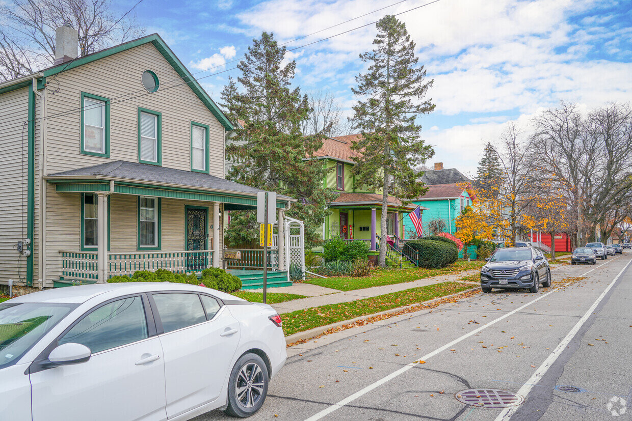 A light gray house with green trim and accents, featuring a covered front porch and a white car parked in front. The neighborhood appears calm and lined with trees.
