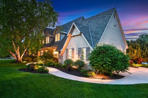 A beautifully landscaped home with well-maintained shrubbery and vibrant green grass, highlighted by landscape lighting in the early evening, illuminating the path leading to the house with a sloped roof and multiple gables.