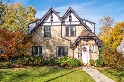 A beautiful Tudor-style stone house with a steeply pitched roof and decorative half-timbering. The home features a white arched front door with red brick trim, framed by a well-maintained lawn and fall-colored trees, giving it a cozy, storybook appearance.