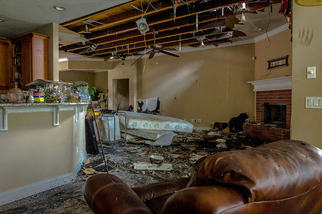 A photo of a fire-damaged living room with visible soot and debris. The ceiling has collapsed partially, exposing burnt wooden beams and damaged drywall. The room is filled with destroyed furniture, including a charred couch, a ruined mattress, and various household items scattered across the floor. The aftermath of the fire has left the space uninhabitable.