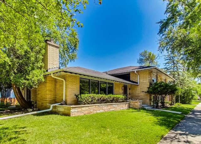 A single-story, mid-century modern brick house with a large green lawn and mature trees. The home has a sloped roof, large front windows, and a chimney, giving it a classic suburban look.
