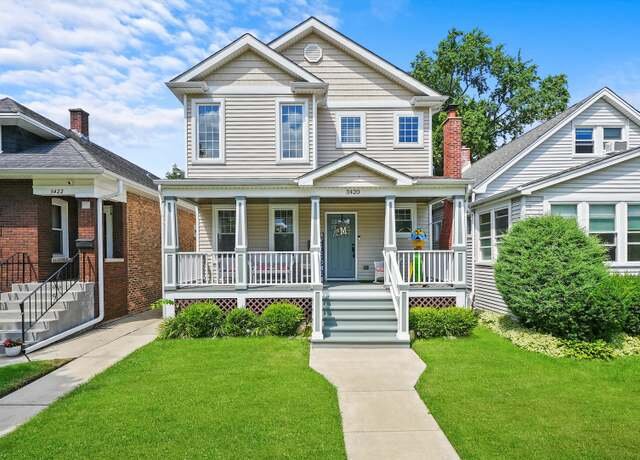 A well-maintained single-family home with a light gray exterior and white trim. The house has a cozy front porch with railings and a few steps leading up, surrounded by lush greenery. The front lawn is neatly landscaped, enhancing the curb appeal of this charming residential property.