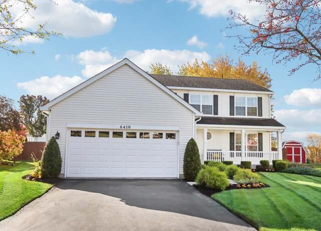  A cozy two-story white house with black shutters, a welcoming front porch, and a paved driveway leading to a double garage. The front yard is landscaped with green shrubs and a well-kept lawn.
