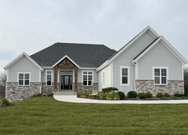 A modern ranch-style home with a stone-accented exterior, a dark gray roof, and light gray siding. The house features large windows, a welcoming front entrance with a wooden beam accent, and a spacious, well-maintained lawn in front.
