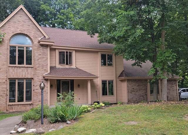 A spacious two-story brick and wood house with a combination of light brown brickwork and wooden paneling. The home is surrounded by tall trees, creating a peaceful, shaded setting, with a front walkway leading to the main entrance.