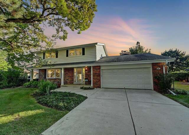 A two-story suburban home with a front-facing garage. The house is built with a mix of red brick and white siding. The driveway leads up to the garage, and the lawn is lush with green grass, surrounded by trees. The sky above displays a colorful sunset with shades of purple and orange.