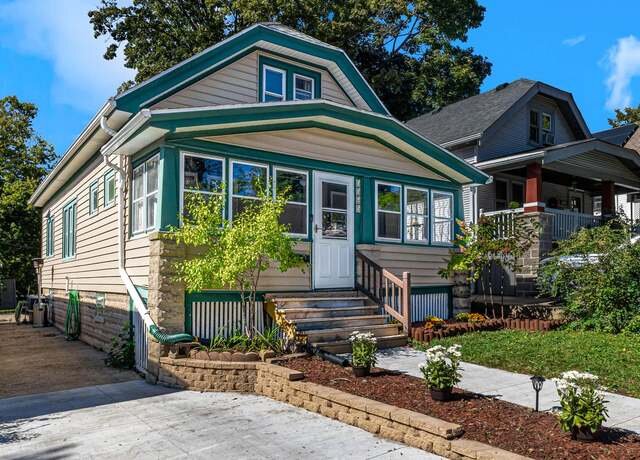 A charming bungalow with green and white trim, featuring a large front porch and a newly landscaped front yard, including flower beds with young plants, giving a welcoming, updated appearance.