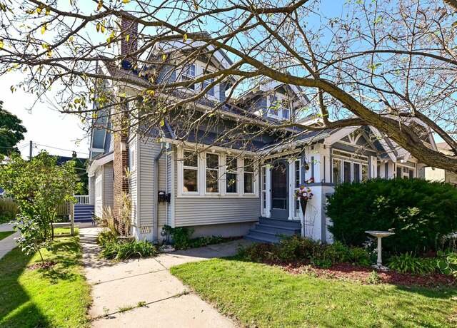 A charming light gray, two-story house with multiple front windows and an inviting porch area surrounded by greenery and mature trees. A lush front yard with a sidewalk leads up to the house, with branches from a nearby tree partially framing the home.