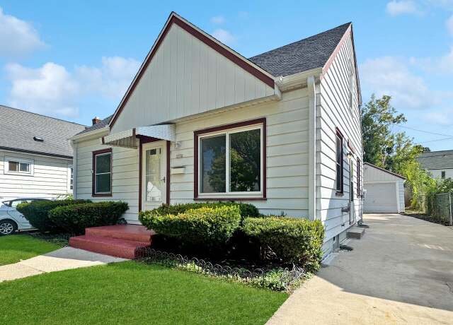 A charming single-family home with a neat front yard, a paved driveway, and an attached garage. The house has a white exterior with red accents and a small front porch.