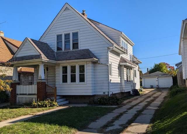 A classic white house with a front porch and a long driveway leading to a detached garage. The roof has gabled architecture, and the house sits under a clear blue sky.