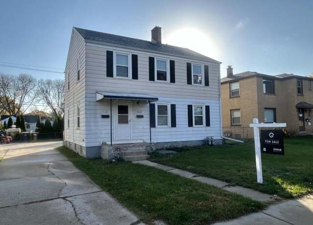 A two-story white house with black shutters on all windows, located on a quiet street with a "For Sale" sign in the yard. The simple design and the sunlight filtering over the roof add a warm touch to this classic American home.