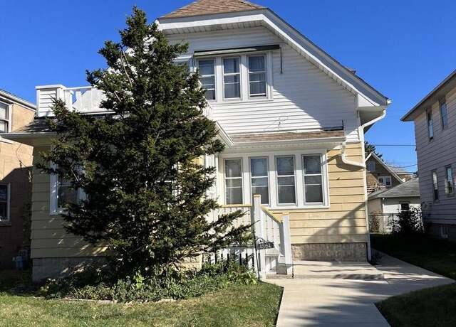 A cozy two-story house with light tan siding and a sloping roof. A tall evergreen tree shades the front yard, with a sidewalk leading up to a small front porch. The house has multiple windows, giving it a bright and welcoming appearance.
