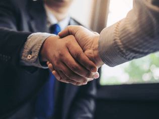 Close-up of two business professionals shaking hands, symbolizing a successful real estate deal. One person is wearing a dark suit, and the other is in a light-colored shirt, with a blurred background of an office setting.