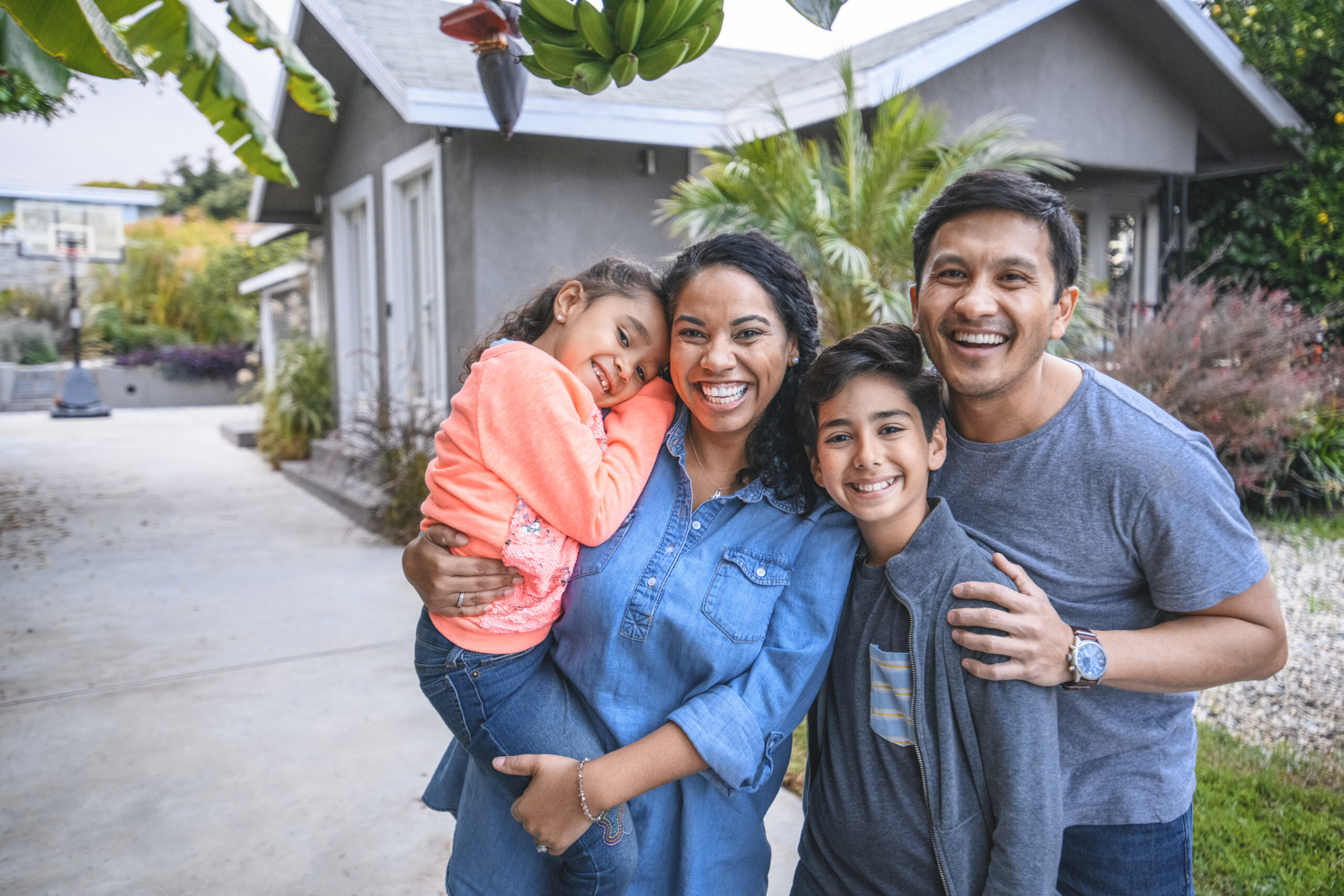  A happy family of four standing together outdoors in front of a gray home. The mother is holding their young daughter in her arms, while the father and their son smile brightly. The scene conveys warmth, happiness, and a sense of togetherness in a comfortable home environment.