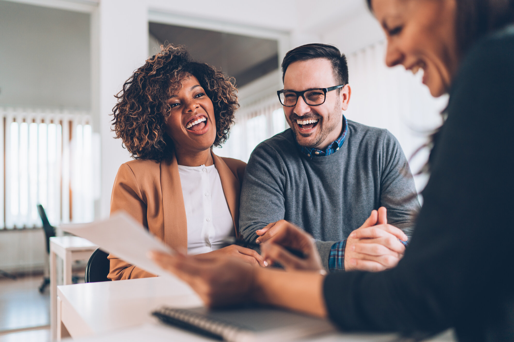 A happy couple sitting at a desk, laughing and engaging in a positive conversation with a professional. They are reviewing documents together, indicating a successful meeting or agreement in a bright, professional office environment.