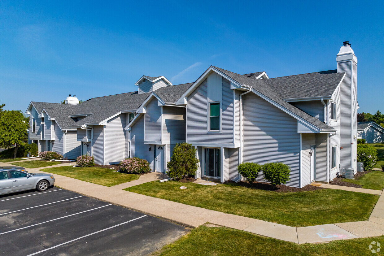 A two-story townhouse-style apartment building with white and gray siding and pitched roofs under a bright blue sky. The area includes parking spots and tidy pathways leading to each entrance, giving a welcoming suburban feel.