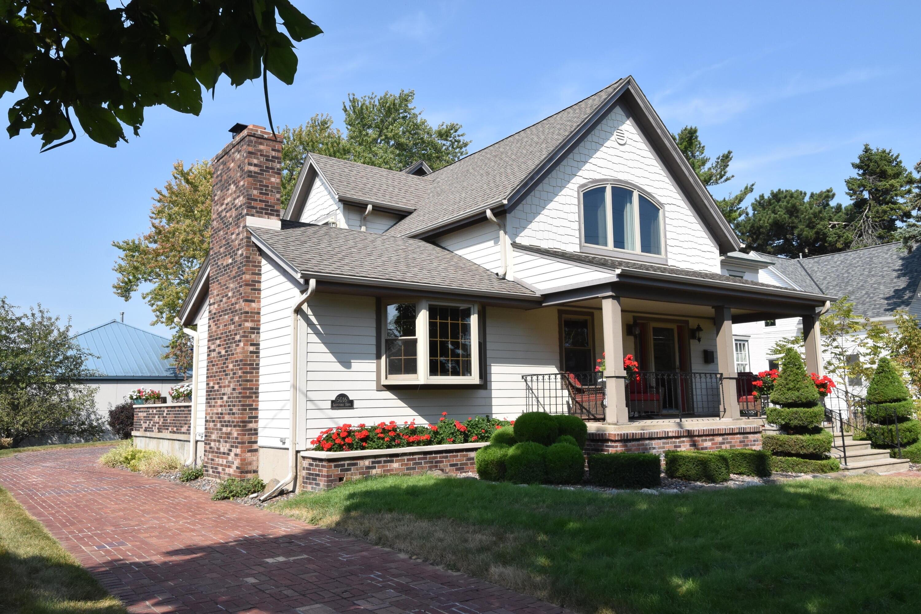 A white suburban home with gray trim and a prominent brick chimney, surrounded by vibrant flower beds and sculpted greenery. A red brick pathway leads to the entrance, adding to the home's inviting appeal.