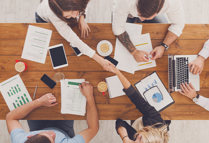  An overhead view of a business meeting with five people sitting around a wooden table. The participants are working on laptops, analyzing documents, and shaking hands in agreement, symbolizing successful collaboration and teamwork.