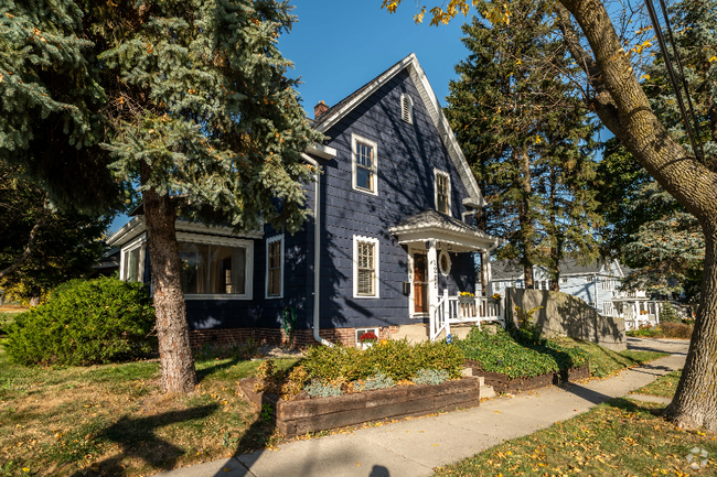 A classic navy-blue, two-story house with white trim accents, giving it a warm, traditional look. The home is nestled among tall trees and has a small white fence surrounding the front porch, complemented by a vibrant front yard garden.