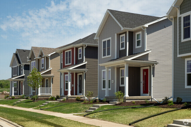 A row of modern two-story suburban homes, each with different siding colors, well-maintained lawns, and small front porches, lining a quiet residential street.