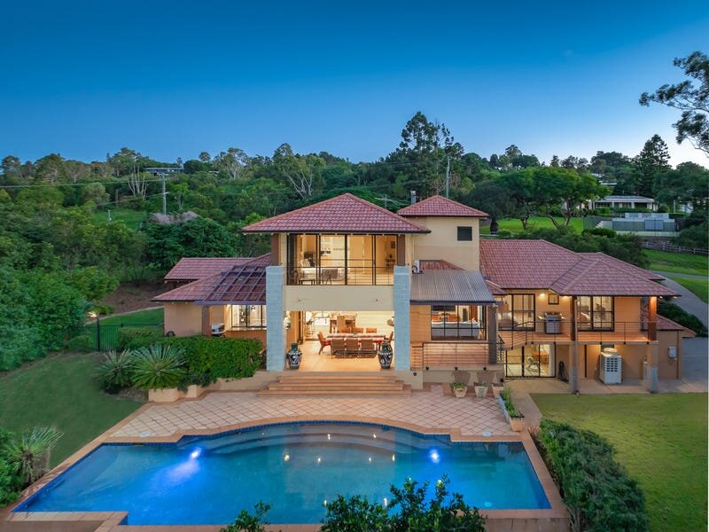 A modern two-story house with a terracotta-tiled roof and large glass windows, illuminated in the evening. The house overlooks a beautifully designed swimming pool and lush garden, with a peaceful hillside setting in the background, making for a serene and upscale atmosphere.